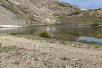 mountains around a lake are covered with snow and rocks and grass around it with small trees and shrubs in the foreground