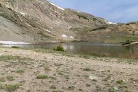 mountains around a lake are covered with snow and rocks and grass around it with small trees and shrubs in the foreground