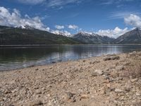 a lake with rocks on the shore and mountains in the background along side it and snow capped hills