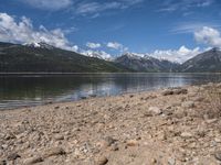 a lake with rocks on the shore and mountains in the background along side it and snow capped hills