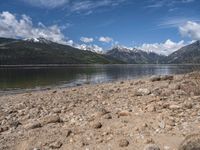 a lake with rocks on the shore and mountains in the background along side it and snow capped hills