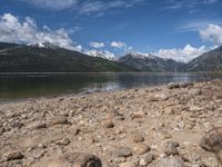 a lake with rocks on the shore and mountains in the background along side it and snow capped hills