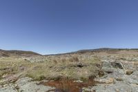 a small pond in the middle of a rocky plain in the background is a field with a mountain and grassy slope