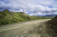 a dirt road leading to mountains in the distance and clouds above it on the left