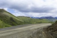 a dirt road leading to mountains in the distance and clouds above it on the left