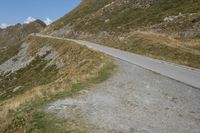 a dirt road that passes between a steep mountain and grassy area on the far side of the road