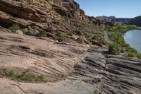 a mountain trail with rock formations above it and a river flowing through it in the foreground