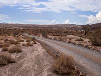 there is a dirt road that runs through a desert landscape with clouds above it and mountains in the distance