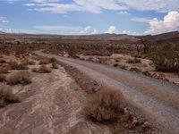 there is a dirt road that runs through a desert landscape with clouds above it and mountains in the distance