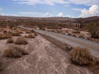 there is a dirt road that runs through a desert landscape with clouds above it and mountains in the distance