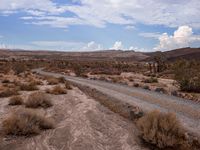 there is a dirt road that runs through a desert landscape with clouds above it and mountains in the distance