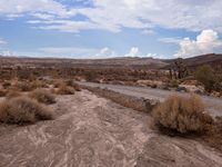 there is a dirt road that runs through a desert landscape with clouds above it and mountains in the distance