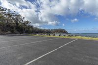 empty parking lot with ocean in background and trees lining side of road and grass to side