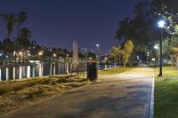 a path with benches and lights along it next to water, in the distance is a city at night