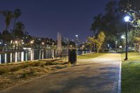 a path with benches and lights along it next to water, in the distance is a city at night