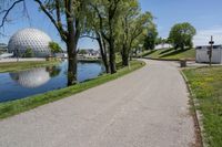 paved road next to grassy field next to river and glass dome in background, with blue sky in front