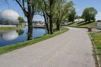 paved road next to grassy field next to river and glass dome in background, with blue sky in front