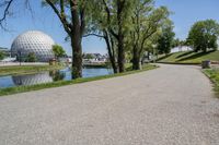 paved road next to grassy field next to river and glass dome in background, with blue sky in front