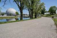 paved road next to grassy field next to river and glass dome in background, with blue sky in front