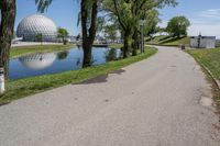 paved road next to grassy field next to river and glass dome in background, with blue sky in front