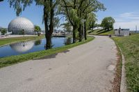 paved road next to grassy field next to river and glass dome in background, with blue sky in front