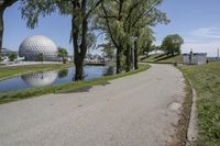 paved road next to grassy field next to river and glass dome in background, with blue sky in front