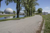 paved road next to grassy field next to river and glass dome in background, with blue sky in front