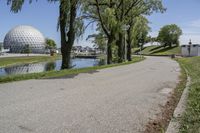 paved road next to grassy field next to river and glass dome in background, with blue sky in front