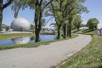 paved road next to grassy field next to river and glass dome in background, with blue sky in front