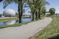 paved road next to grassy field next to river and glass dome in background, with blue sky in front