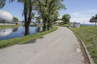 paved road next to grassy field next to river and glass dome in background, with blue sky in front