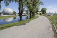 paved road next to grassy field next to river and glass dome in background, with blue sky in front