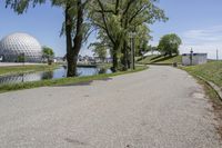 paved road next to grassy field next to river and glass dome in background, with blue sky in front