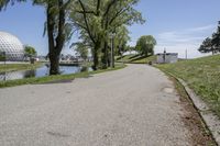 paved road next to grassy field next to river and glass dome in background, with blue sky in front