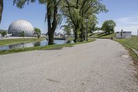 paved road next to grassy field next to river and glass dome in background, with blue sky in front