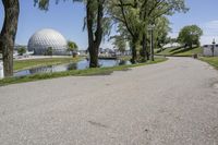 paved road next to grassy field next to river and glass dome in background, with blue sky in front