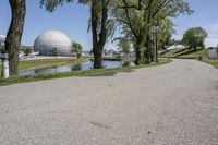 paved road next to grassy field next to river and glass dome in background, with blue sky in front