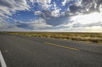 a paved street leads to the horizon of a prairie with clouds overhead and grass all around