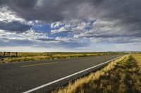 a paved street leads to the horizon of a prairie with clouds overhead and grass all around