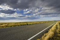 a paved street leads to the horizon of a prairie with clouds overhead and grass all around