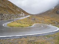 a curve in the road beside a mountain side with low clouds around it to be viewed from above