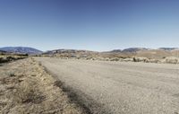 a road leading into an empty area, with grass, dirt and bushes, in the distance are mountains