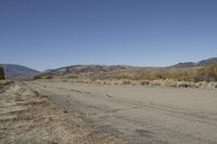 a road leading into an empty area, with grass, dirt and bushes, in the distance are mountains