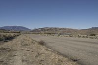 a road leading into an empty area, with grass, dirt and bushes, in the distance are mountains