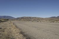 a road leading into an empty area, with grass, dirt and bushes, in the distance are mountains
