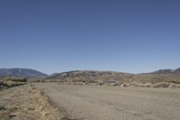 a road leading into an empty area, with grass, dirt and bushes, in the distance are mountains