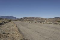 a road leading into an empty area, with grass, dirt and bushes, in the distance are mountains