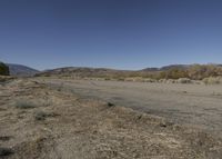 a road leading into an empty area, with grass, dirt and bushes, in the distance are mountains