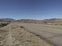 a road leading into an empty area, with grass, dirt and bushes, in the distance are mountains