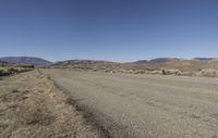 a road leading into an empty area, with grass, dirt and bushes, in the distance are mountains